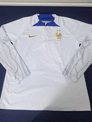Vit Nike Frankrike träningströja - Snygg långärmad träningströja från Nike i vitt med blå detaljer och guldiga loggor. Tröjan har halv dragkedja, Frankrikes landslagsemblem och diskreta mönster på ärmarna. Perfekt för fotbollsträning eller sportiga dagar.