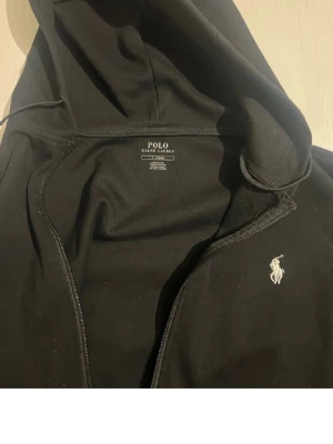 Svart hoodie från Polo Ralph Lauren - Svart hoodie med dragkedja från Polo Ralph Lauren. Klassisk design med vit broderad logga på bröstet och huva med dragsko. Perfekt för en chill och stilren look. Mjukt material som känns skönt mot huden.
