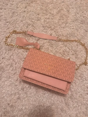 Rosa axelväska från Michael Kors - Säljer en snygg rosa axelväska från Michael Kors med guldiga detaljer och kedjerem. Väskan har MK-logga över hela ytan och ett stilrent lock. Tillverkad i konstläder och textil, perfekt för dig som gillar trendiga accessoarer.