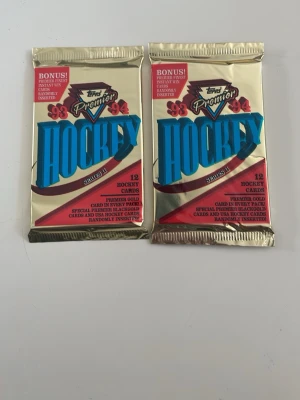 Hockeykortpaket 93-94 Topps Premier - Två oöppnade paket hockeykort från Topps Premier säsongen 1993-94, Series II. Varje paket innehåller 12 kort. Förpackningarna är guldfärgade med blå och röda detaljer. Perfekt för samlare eller hockeyfans som gillar nostalgi.
