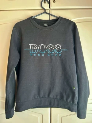 Svart stickad tröja från Hugo Boss - Cool svart stickad tröja från Hugo Boss med stort broderat BOSS-logo i vitt och blått på bröstet. Rund halsringning och ribbade muddar vid ärmslut och nederkant. Perfekt för dig som gillar stilrena och trendiga plagg.