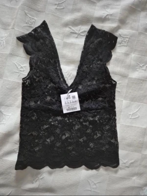 Svart spetslinne från Pull&Bear - Supersnyggt svart spetslinne från Pull&Bear med djup v-ringning och scallop-detaljer vid axlarna. Linnet är ärmlöst och har ett feminint spetsmönster över hela plagget. Perfekt att styla med jeans eller kjol för en trendig look. Lappen är inte kvar