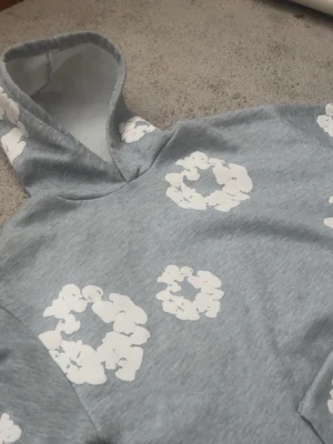  Denim Tears  - Snygg grå hoodie från Denim Tears med vita blommönster tryckta över hela tröjan. Hoodien har en klassisk huva och är gjord i mjukt bomullsmaterial. Perfekt för dig som gillar streetwear och vill sticka ut lite extra.