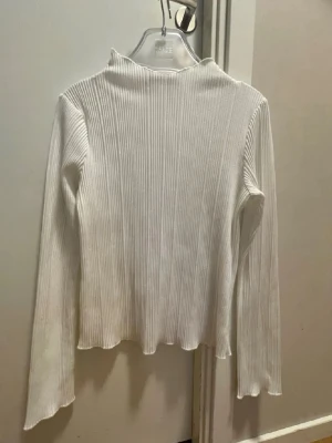 Vit ribbad polotopp från Gina  - Säljer en vit ribbad polotopp med lång ärm och hög hals. Toppen har en smal passform och är tillverkad i ett stretchigt material som sitter snyggt på kroppen. Perfekt att styla med jeans eller kjol för en clean look.
