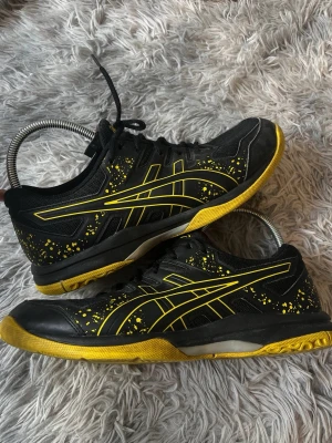 Svarta och gula Asics sneakers - Snygga svarta sneakers från Asics med gula detaljer och stänk, samt gula sulor. Skorna har klassisk Asics-logga på sidorna och är tillverkade i en mix av mesh och syntetmaterial. Perfekta för dig som gillar sportig stil och vill sticka ut med färg.