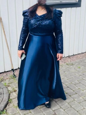 Marinblå långklänning med paljetter - Elegant marinblå långklänning med glittrig paljettöverdel och långa ärmar. Kjolen är i ett glansigt material med vacker fall. Perfekt för speciella tillfällen.