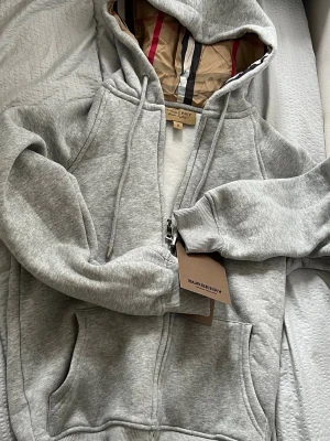 Burberry zip-hoodie grå - Snygg grå hoodie från Burberry med dragkedja, huva med dragsko och klassiskt rutigt foder i huvan. Tillverkad i mjuk bomullsblandning och har ribbade muddar samt två stora fickor framtill. Perfekt för dig som gillar stilrena och exklusiva plagg.