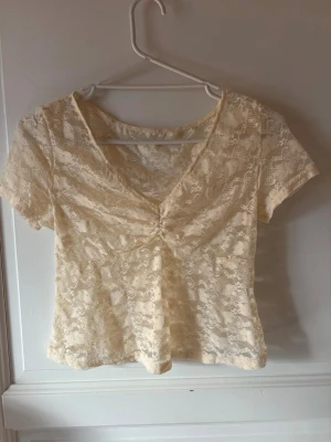 Beige spetsblus med v-ringning - Säljer en beige blus med v-ringning och korta ärmar. Blusen är helt i spets och har en lätt croppad passform med snygg rynkning framtill. 