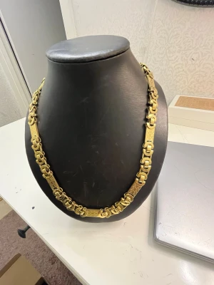 Guldfärgat kedjehalsband med mönster - Säljer ett chunky halsband i guldfärg med unika, rektangulära länkar och detaljerat mönster. Halsbandet har en robust och iögonfallande design som verkligen sticker ut. Perfekt för dig som gillar statement-smycken och vill ha något som syns.