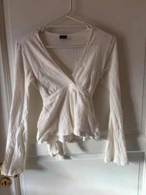 Vit omlottopp med v-ringning - Säljer en vit omlottopp med v-ringning och långa ärmar. Toppen har en lätt strukturerad yta och knytband i midjan för en snygg passform. Perfekt att styla med jeans eller kjol.