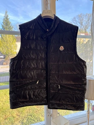 Moncler gui väst - Snygg svart dunväst från Moncler med hög krage och dragkedja framtill. Västen har två snedställda fickor med dragkedja och Moncler-logga på bröstet. Tillverkad i glansigt material och fylld med dun för extra värme.storlek 6 passar dig som bär normal xl/L i kläder kommer med den klassiska påsen till gui väst men även triangeln använd endast 3 gånger som ny