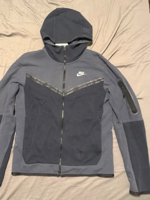 Nike Tech Fleece hoodie mörkblå/silver - Nike Tech Fleece hoodie i mörkgrått med hel dragkedja, huva och silver Nike-logga på bröstet. Snygg V-design framtill och ficka med dragkedja på ärmen. Tillverkad i mjukt och lätt fleecematerial, perfekt för en sportig och avslappnad stil. Pris kan diskuteras!!