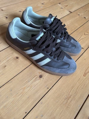 Adidas Samba sneakers brun/grå - Adidas Samba sneakers i brunt läder med grå mockadetaljer och klassiska vita ränder på sidorna. Gummisula och svart snörning. Ikonisk retrostil som funkar till många outfits.