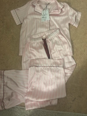 Rosa satinpyjamas från Gina Tricot - Snygg pyjamas i satin från Gina Tricot i ljusrosa med diskreta ränder och mörkare kantband. Setet består av kortärmad skjorta med krage och matchande byxor med resår i midjan. Perfekt för mysiga hemmakvällar.