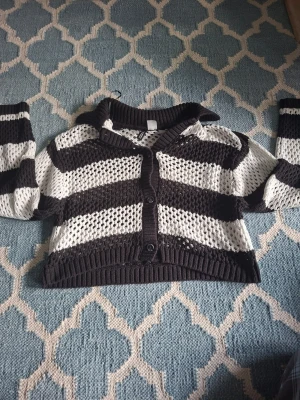 Randig stickad cropped cardigan - Säljer en svartvit stickad cropped cardigan med breda ränder och stora hålstickade partier. Tröjan har långa ärmar, ribbade muddar och knappar framtill. Perfekt för lager på lager och en cool, avslappnad stil.