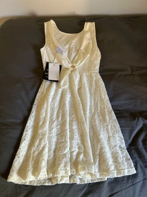 Vit spetsklänning med v-ringning - Superfin vit klänning i spets med v-ringning och ärmlös design. Klänningen har en lätt utställd kjol och en dekorativ knapp framtill. Perfekt för student eller avslutning💞💞💞