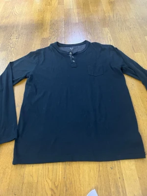 Svart långärmad topp med knappar - En svart långärmad topp med rund halsringning, knappdetaljer framtill och en liten bröstficka. Tillverkad i mjuk bomull som känns skön mot huden. Enkel och clean design som passar till det mesta.