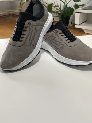 Grå sneakers med svart detalj - Snygga sneakers i grått med svart hälparti och vita sulor. Skorna har snörning och en stilren, modern design med diskret perforerat mönster på sidorna. Perfekta för dig som gillar en clean look och vill ha bekväma skor till vardags.helt ny 