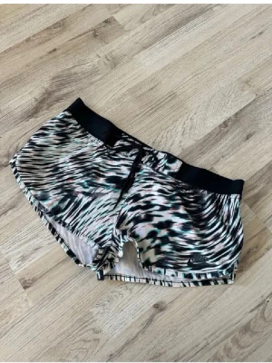 Nike träningsshorts med mönster - Nike träningsshorts med svart resår och dragsko i midjan. Shortsen har ett coolt abstrakt mönster i svart, vitt, grönt och rosa. Lätta och luftiga, perfekta för löpning eller gym. Nike-logga på benet.