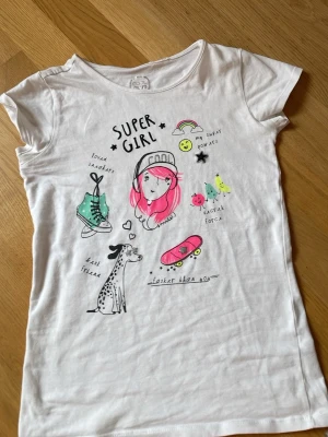 Vit t-shirt med Super Girl-tryck - Vit t-shirt med färgglatt Super Girl-tryck framtill, motiv som sneakers, skateboard, dalmatiner och regnbåge. Rund halsringning och korta ärmar. Skön och mjuk bomullskvalitet, perfekt för en avslappnad stil.