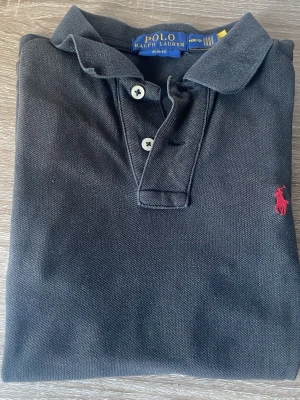 Svart pikétröja från Ralph Lauren - Klassisk svart pikétröja från Ralph Lauren med krage, knappar och den ikoniska röda logotypen broderad på bröstet. Tillverkad i bomull för en bekväm och stilren look. Perfekt till jeans eller shorts.