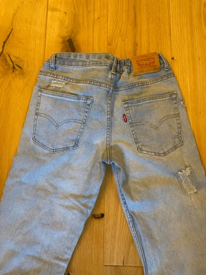 Levi's ljusblå raka jeans - Säljer ett par ljusblå raka jeans från Levi's med klassisk femficksdesign och slitna detaljer på ben och fickor. Jeansen har en avslappnad passform och är perfekta för en cool vardagslook. Levi's-logga på bakfickan och läderpatch i midjan.