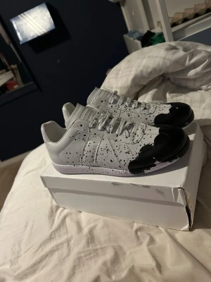 Vita sneakers med svart splash-design - Unika vita sneakers med svart färgstänk och svartmålade tår. Skorna har klassisk låg profil, vita snören och ovandel i syntetmaterial. Perfekta för dig som vill sticka ut med en kreativ och modern look. Kom med bud eller funderingar aldrig använda strl 42/43
