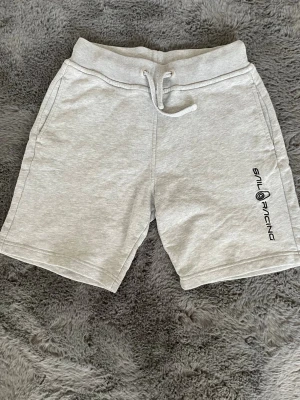 Grå Sail Racing shorts med snörning - Snygga ljusgrå shorts från Sail Racing med snörning i midjan och tryckt logga på ena benet. Tillverkade i mjuk bomullsmix och har fickor fram samt en bakficka. Perfekta för chill dagar eller sommarens äventyr.