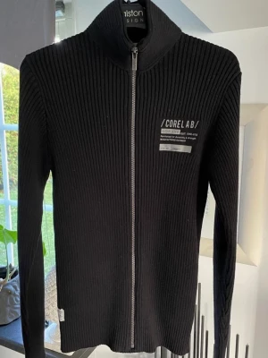 Svart ribbad ziptröja Jack & Jones - Svart ribbstickad ziptröja från Jack & Jones Core Lab med hög krage och tryckt logga på bröstet. Tröjan har hel dragkedja framtill och är tillverkad i ett stretchigt material som sitter snyggt. Perfekt för en stilren och modern look.