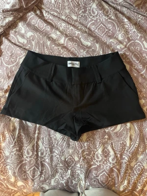 Svarta shorts från Princess Polly - Svarta shorts från Princess Polly med klassisk design och bälteshällor. De har fickor fram och bak, och är sydda i ett slätt material som ger en stilren look. Perfekta för en trendig och enkel outfit. Lågmidjade kostym shorts.