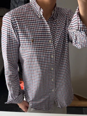 Rutig skjorta från Ralph Lauren - Klassisk rutig skjorta från Ralph Lauren i vitt, blått och rött. Skjortan har lång ärm, knappar framtill och en broderad logga på bröstet. Tillverkad i mjuk bomull och har en normal passform. Perfekt för en clean och stilren look. Lite missfärgad i kragen och armhålorna, därav priset. Annars bra skick 