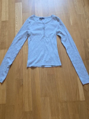 Ljusgrå långärmad topp med knappar - Ljusgrå långärmad topp från new yorker med rund halsringning och dekorativa knappar framtill. Slim fit och mjukt bomullsmaterial som känns skönt mot huden. Perfekt att matcha med jeans eller kjol för en enkel och clean look.