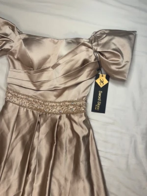 Balklänning champagnefärgad långklänning i satin oandvänd - Elegant oandvänd långklänning i champagnefärgad satin från Sword Stone. Klänningen har offshoulder-ärmar, och ett dekorerat midjeband med pärlor och paljetter. Originellt köpt för 3999kr. Fick tyvärr aldrig tillfälle att andvända den. Den har även en slits vid benet men också en underkjol. Perfekt inför en bal eller fester! 💕