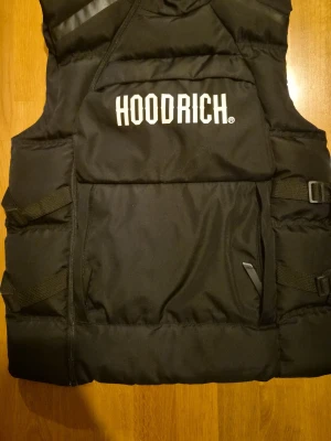 Svart dunväst från Hoodrich - Svart dunväst från Hoodrich med stor vit logga på bröstet. Västen har quiltad design, justerbara band i sidorna och dragkedja framtill. Perfekt för lager-på-lager och streetwear-stil.