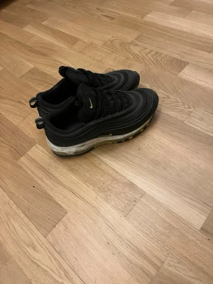 Nike Air Max 97 svarta sneakers - Svarta Nike Air Max 97 sneakers med vågformade detaljer och synlig Air-sula. Skorna har gula detaljer på loggan och sulan, samt dragflikar bak och fram. Ovandelen är i textil och syntet, med en sportig och modern look.