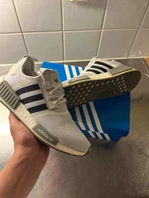 Adidas NMD R1 vita sneakers - Fräscha Adidas NMD R1 sneakers i vitt mesh med svarta ränder och grå detaljer. Skorna har en chunky vit Boost-sula och klassiska NMD-block på sidorna. Perfekta för en sportig och stilren look. Levereras med originalkartong.