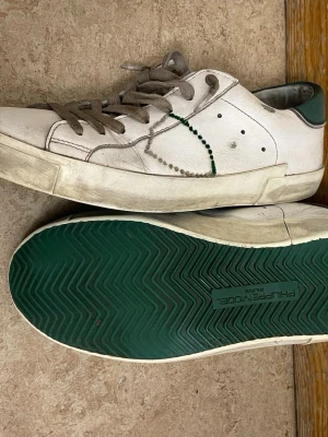 Vita låga sneakers från Philippe Model - Säljer ett par vita sneakers från Philippe Model med gröna detaljer och grön sula. Skorna har grå snörning och klassisk låg siluett. Materialet är läder och designen har subtila sömmar och prickdetaljer på sidan.