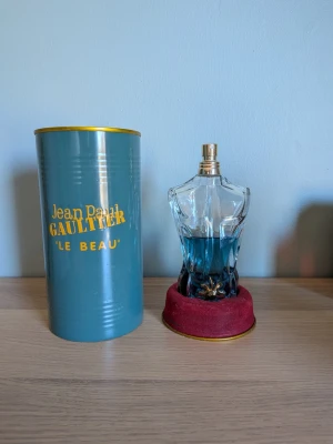 Jean Paul Gaultier Le Beau parfym 10ml - Jean Paul Gaultier Le Beau parfym i en unik flaska formad som en manlig torso med blått innehåll. Kommer med originalförpackning i metall med guldfärgade detaljer och text. Flaskan står på en röd sammetsliknande bas med guldkant.