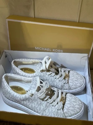 Michael Kors vita mönstrade sneakers - Snygga vita sneakers från Michael Kors med diskret logomönster över hela skon. Skorna har vita snören med guldfärgade detaljer och en klassisk låg siluett. Tillverkade i textilmaterial med gummisula. Perfekta för en trendig och stilren look.