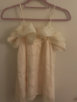 Ljus blus från Elli White - Supersöt ljus blus från Elli White med axelbandslös design och stora volangdetaljer upptill. Blusen har en flowy form och är tillverkad i ett lätt, skimrande material som ger en exklusiv känsla. Perfekt för dig som vill sticka ut med en unik look.