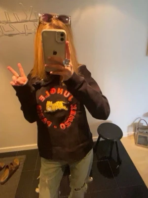 Svart Kenzo hoodie med tigertryck - Cool svart hoodie från Kenzo med färgglatt tigertryck och texten 'Jungle Kenzo Paris'  på bröstet. Unik volangdetalj över axeln och halsen. Perfekt för dig som vill sticka ut med en trendig och exklusiv look. Super bra skick! Knappt använd. 