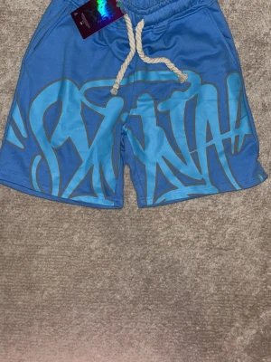 Blå shorts från OYAKUCKABIES - Snygga blå shorts från OYAKUCKABIES med stort grafiskt tryck framtill och vit snörning i midjan. Perfekta för sommaren med en avslappnad vibe och cool streetstil. Shortsen har fickor på sidorna och elastisk midja för skön passform.