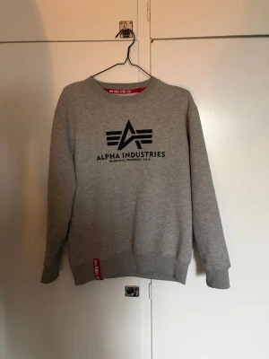 Grå sweatshirt från Alpha Industries - Snygg grå sweatshirt från Alpha Industries med svart logotryck på bröstet, väldigt fint skick ingen syn av användning 