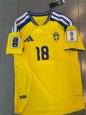 Svensk landslagströja Adidas gul - Gul svensk landslagströja från Adidas med blå detaljer och nummer 18 på bröstet. Tröjan har FIFA och VM patchar på ärmarna, samt Sveriges emblem på vänster bröst. Materialet är lätt och andas, ClimaCool-teknologi.