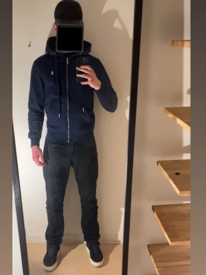 Mörkblå hoodie från Polo Ralph Lauren - Snygg mörkblå hoodie från Polo Ralph Lauren med dragkedja framtill och klassisk huva. Ikoniska loggan broderad på bröstet. Mjukt material och ribbade muddar vid ärmslut och nederkant. Perfekt för en avslappnad och stilren look.