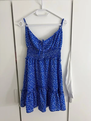 Blå sommarklänning med vita blommor - Supersöt blå klänning med vita småblommiga mönster och smockad midja. Klänningen har tunna axelband, volangkant nertill och en luftig, kort modell som passar perfekt till varma dagar. Materialet känns lätt och flowy.