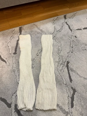 Vita ribbade benvärmare - Säljer ett par vita ribbade benvärmare i mjukt stickat material. Perfekta att dra över leggings eller jeans för en extra touch till outfiten. De är långa och har en klassisk ribbad struktur som ger en snygg och enkel look.