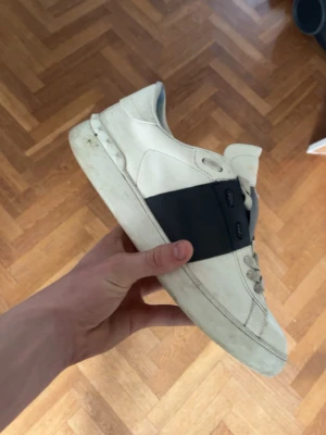 Vita Vale open sneakers med svart band - Snygga vita sneakers från Valetino med ett brett svart band över mitten. Skorna har klassisk låg profil och är tillverkade i läder med vita snören och diskreta detaljer. Perfekta för dig som gillar stilrena och exklusiva sneakers.