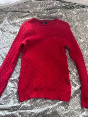 Röd kabelstickad tröja från Gant - Snygg röd kabelstickad tröja från Gant med rund halsringning och diskret logga på bröstet. Tröjan har långa ärmar och ribbade muddar vid ärmslut och nederkant. Perfekt för dig som gillar klassisk och stilren design. Är liten i storleken så passar bättre på folk som har small i vanliga fall