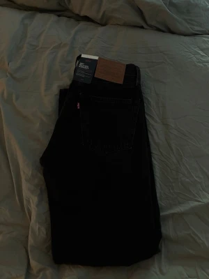 Svarta Levi's 555 jeans - Säljer ett par klassiska svarta Levi's 555 jeans med raka ben och femficksmodell. Jeansen har den ikoniska läderlappen bak i midjan och Levi's röda etikett på bakfickan. Tillverkade i kraftig denim som ger en tidlös look. Helt nya och oanvända.